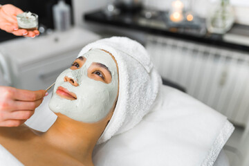 Wall Mural - Deep purifying procedure at SPA. Portrait of beautiful positive smiling Asian woman in her 20s with white towel on her hair lying down on SPA bed while experienced cosmetician is putting clay mask on