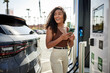 © Cavan Images - Happy young woman with phone at electric vehicle charging station
