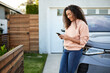 © Cavan Images - Smiling woman using smart phone leaning on electric car in front yard