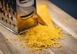 A heap of grated cheddar cheese next to a grater and a cheese chunk on wooden cutting board