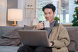 © wattana - Smiling handsome asian man sitting on floor in living room and using laptop computer.