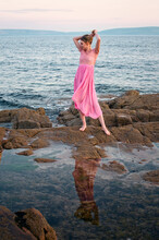Beautiful Young Caucasian Girl In Pink Dress Standing On The Rocky Coast Reflected In Water At Barna, Galway, Ireland 