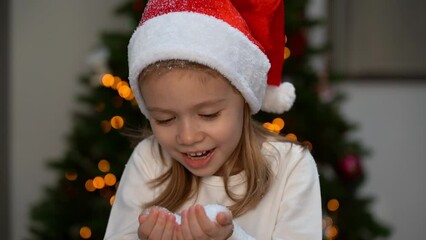 Wall Mural - Little girl fun blows snow into camera on background of Christmas tree at home and laugh. Happy child plays with artificial snow. New year and christmas celebration