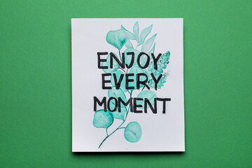 Wall Mural - Card with beautiful phrase Enjoy Every Moment on green background, top view