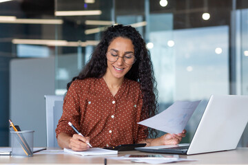 Wall Mural - latin american businesswoman working inside office with documents and laptop, worker paperwork calculates financial indicators smiling and happy with success and results of achievement and work