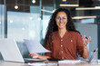 © Liubomir - Portrait of happy and successful hispanic woman, businesswoman smiling and looking at camera holding contracts and invoices, working inside office with laptop on paper work.