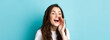 © Mix and Match Studio - Close up portrait of cheerful young woman calling someone, holding hand near opened mouth while shouting, searching for person, speaking loud, standing over blue background