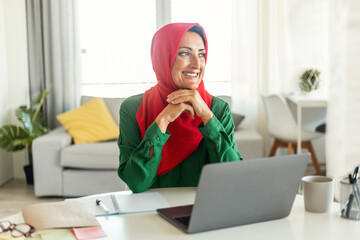 Wall Mural - Positive islamic woman in hijab sitting at desk with laptop at home and thinking about something and smiling