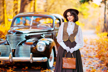 Beautiful female in vintage dress, lace blouse and  veil standing near retro brown car on autumn background. Elegant lady  with gloves on nature.Warm art work.