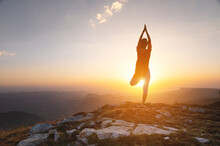 Silhouette Of A Woman In A Tree Pose, Yoga Asana, Stands Near A Cliff In The Mountains Against The Backdrop Of A Sunset With A Clear Sky