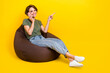 © deagreez - Full length photo of nice positive woman brown hair talking phone sit on pouf directing empty space isolated on yellow color background