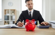 © Studio Romantic - Business and finance. Piggy bank on table next to serious man in suit who uses calculator to make calculations. Close up of piggy bank in form of red pig in glasses on background of businessman.