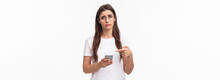 Communication, Technology And Lifestyle Concept. Waist-up Potrait Of Sad And Gloomy Young Uneasy Caucasian Woman In Glasses, Frowning And Pointing At Mobile Phone With Regret Or Sadness
