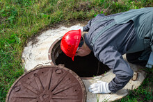 A Male Plumber Opened The Hatch Of A Water Well And Looks Inside. Inspection Of Water Pipes And Meters