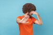 © khosrork - Stop, I don't want to see at this. Portrait of man with Afro hairstyle wearing orange T-shirt covering his face and showing stop hand gesture. Indoor studio shot isolated on blue background.