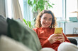 © insta_photos - Smiling young pretty woman sitting on couch using apps on cell phone technology, happy lady holding smartphone in hands, looking at camera, relaxing on sofa with cellphone checking cellular device.