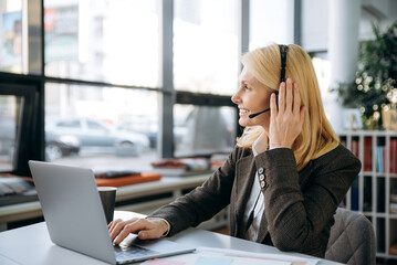 Wall Mural - Concentrated business woman sitting at the desk, wearing headset, communicate with colleagues. Smiling female call center operator is consulting a clients, support service concept