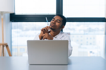 Poster - Portrait of serious black male doctor in white uniform looking to syringe with vaccine in hand sitting at table with laptop in clinic on background of window. Concept of vaccination, immunization.