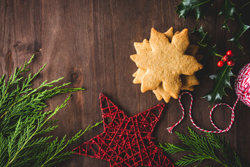 galletas de Navidad en forma de estrella o copo de nieve, junto a ramas de ciprés y acebo, adorno de estrella y cordón rojo y blanco, sobre una mesa de madera