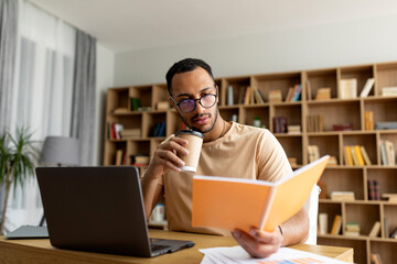 Wall Mural - Arab man studying online from home, reading notebook and drinking coffee, sitting at desk with laptop pc, free space