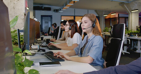 caucasian beautiful cheerful woman in headset working at computer and talking with client in office.