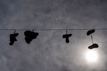 A Dramatic Sky With White Clouds Behind Which Is The Sun, But A Black Rope Is Stretched Across It, On Which A Few Pairs Of Shoes Are Hanging