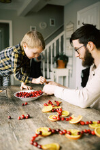 Father And Son Making Orange And Cranberry Garlands For Christmas.