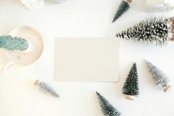 Wall Mural - Christmas card mock up. Empty greeting card and little christmas trees decorations flat lay on rustic white table. Postcard template with space for text. Happy Holidays!