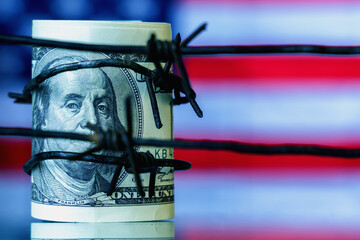 US Dollar currency wrapped in barbed wire against flag of United States as symbol of economic warfare, sanctions and embargo busting concept.  Free copy space for design or text.