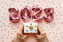 A Person Is Holding A White Gift Box With A Golden Boy And A Gift On A Pink Background. Next To Confetti And Gold And Silver Numbers 2023. Festive Background Of The Concept Of New Year And Christmas.