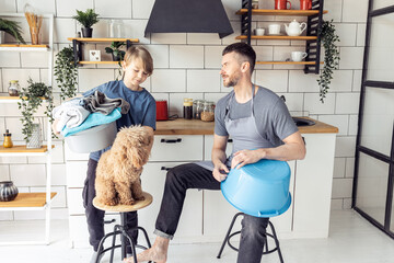 Wall Mural - Handsome father and his teenager son spending quality time together, having fun. Men doing chores, cleaning, sorting laundry in the kitchen at home. Pet dog near them