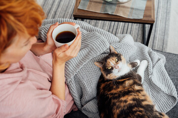 Close-up woman in a plaid drinking hot tea, petting a relaxed cat on the sofa at home. Cozy and comfortable winter or autumn weekends. Pleasant ways to keep warm. Take a break and relax