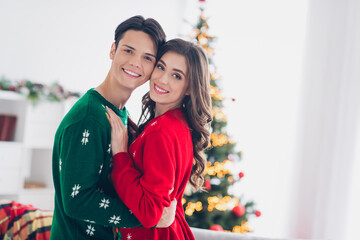 Poster - Photo of young couple together cuddles wear ugly sweaters enjoy spend time together christmastime near fir tree lights illumination indoors