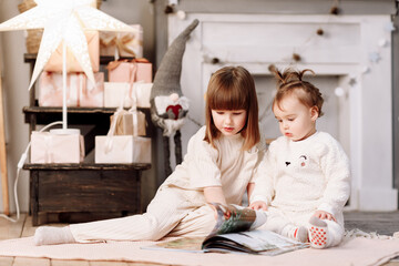 Two happy little child girls sitting on floor and reading a story book together by fireplace in a decorated cozy light living room on winter holiday. Celebrating Xmas and New Year at home with family