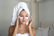 Young woman wearing a towel after showering, pampering the under eye patch for dark circles treatment. Female applying cosmetic products to decrease puffiness under her eyes. Close up, background.