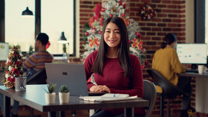 Wall Mural - Portrait of female worker using laptop in festive office filled with winter decorations and ornaments. Woman working on startup during christmas holiday festivity in space with xmas tree.