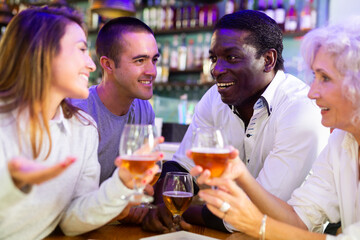 Wall Mural - International group of glad cheerful people toasting with beer, having fun at party in beer pub. Focus on young man