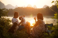 Asian Family Of Three Having A Picnic By The River At Sunset