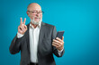 © kinovo - Video call by phone. Handsome elderly man greets a friend using a mobile phone in a classic suit on a blue background. studio portrait