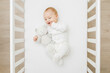 Adorable happy baby boy in white bodysuit hugging teddy bear and lying down on back on mattress in crib at home room. 6 months old infant playing with first friend. Closeup. Top down view.
