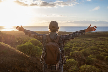 Back view of senior caucasian woman with hat and casual clothing standing with open arms looking at the horizon over sea. Elderly lady enjoying nature and freedom admiring landscape at sunset light