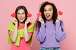 © ViDi Studio - Young two friends happy surprised women 20s wears green purple shirts together hold in hand red paper hearts stickers look camera isolated on pastel plain light pink color background studio portrait.