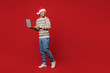 © ViDi Studio - Full body merry young IT man wears warm cozy Christmas sweater Santa hat posing hold use work on laptop pc computer isolated on plain red background. Happy New Year 2023 celebration holiday concept.