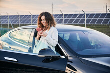 Pretty Girl Looking At Her Smartphone While Standing Next To Black Auto With Opened Side Door. Smiling Lady With Phone In Hands Leaning Against Side Of Open Car. On The Backdrop Solar Power Plant.