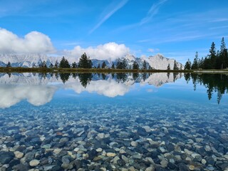 Canvas Print - Small rocks under shallow lake near shore with the reflection of cloudy sky and distant mountains