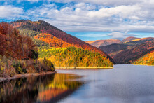Scenic View Of Autumn Colored Forest Reflecting To Alpine Lake In Transylvanian Alps During Warm October Light Against Dramatic Sky