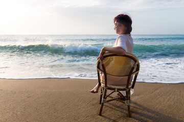 Wall Mural - An adult woman on the beach is sitting on a chair looking at the sea waves.