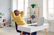 © Studio Romantic - Smiling young woman sitting in front of laptop with arms behind head. Portrait of happy African American girl relaxing while sitting at desk at cozy interior of home or office