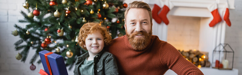 cheerful and redhead father and son looking at camera in living room with Christmas tree and fireplace, banner