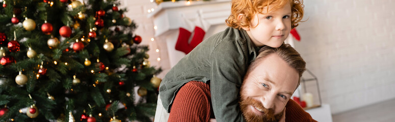 Wall Mural - happy bearded man piggybacking redhead son near blurred Christmas tree, banner